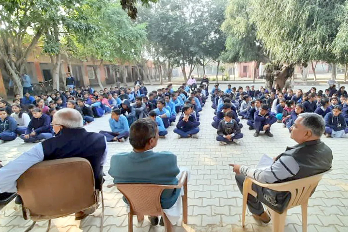 Schoolchildren seated in rows in a school courtyard, with volunteers watching